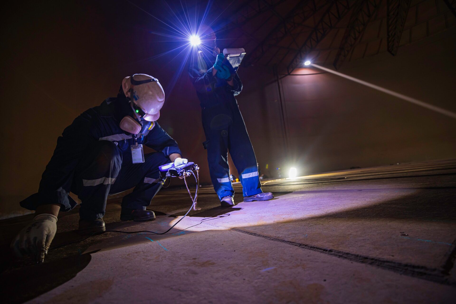 Two confined space standby personnel conducting safety checks, with one kneeling and the other standing and using a light source, highlighting the responsibilities of standby workers in confined space environments.
