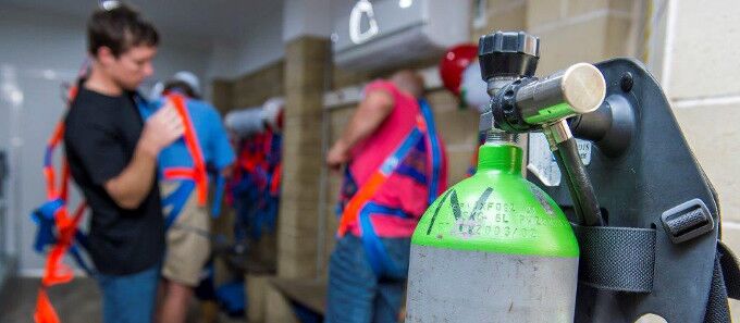 A close-up of a breathing apparatus tank in a preparation area, with individuals in the background getting ready for service and inspection, highlighting the importance of compliant breathing apparatus checks.