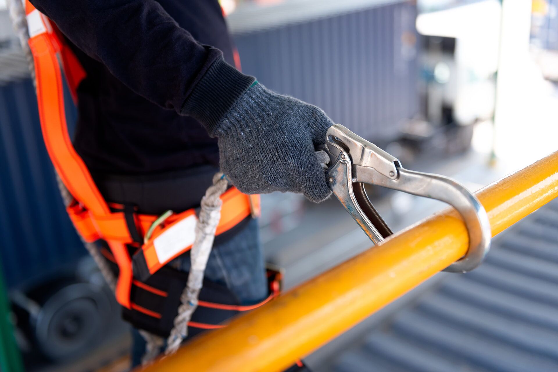 Person wearing a safety harness and gloves, holding a carabiner on a yellow railing while performing a working at height risk assessment.