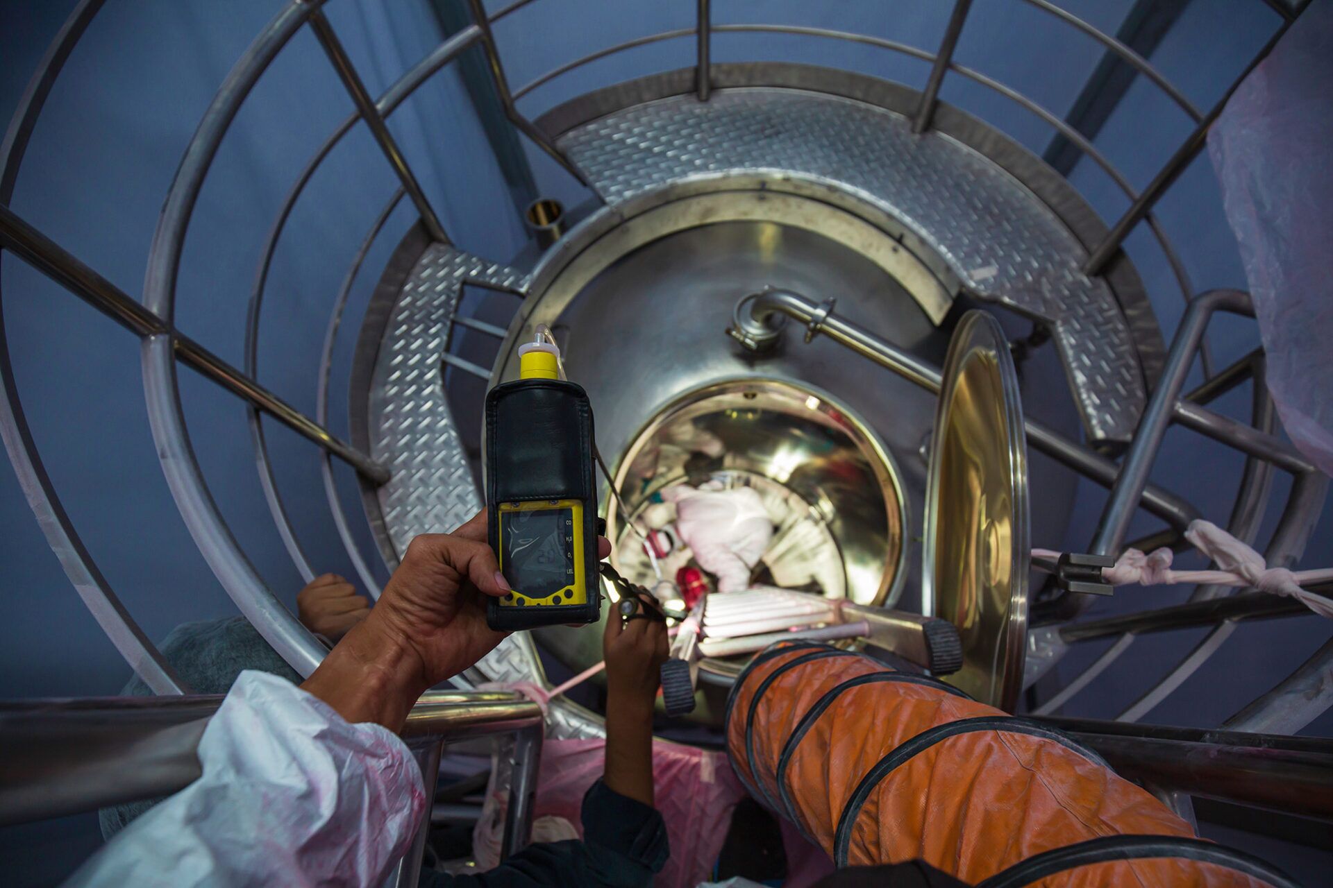 Hand holding a portable oxygen monitor while looking down into a confined space, emphasizing the importance of checking oxygen levels in confined environments.