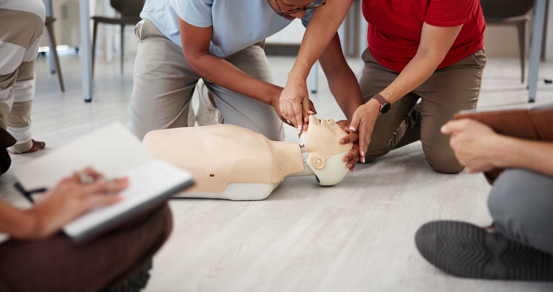 Two participants practicing cardiopulmonary resuscitation (CPR) techniques on a training mannequin during a CPR training course in Sydney. A notebook is visible in the foreground, indicating an interactive learning environment.