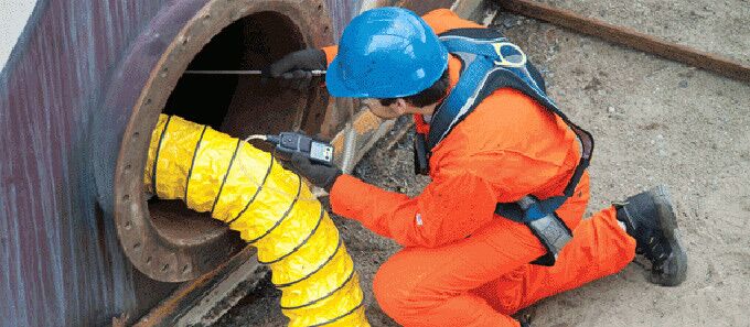 Worker in an orange jumpsuit and blue helmet conducting a safety assessment in a confined space using a gas detector and ventilation duct for confined space rescue plans.